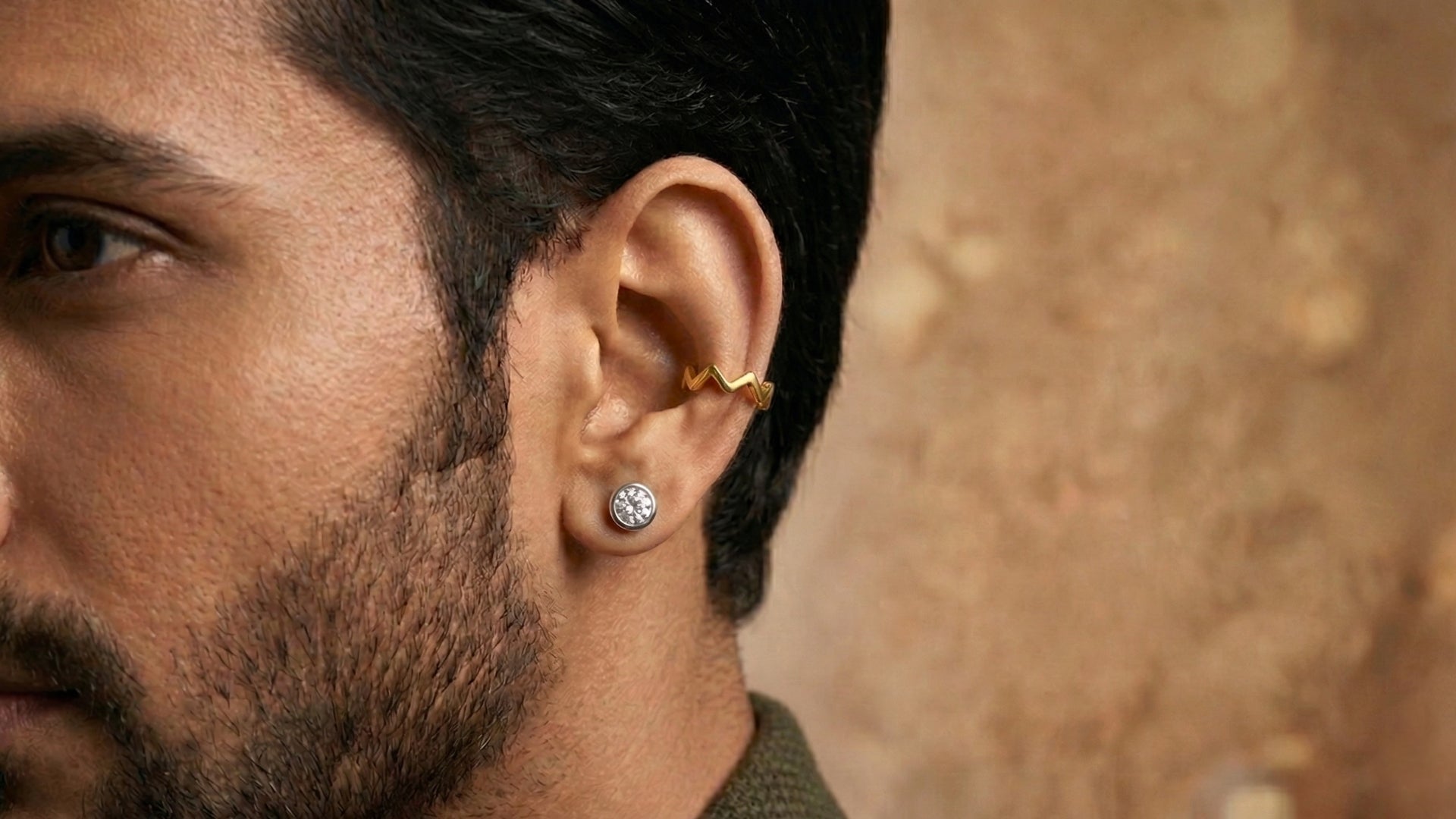 Close-up of a man with a beard wearing a round diamond stud earring on the lobe and a gold zigzag ear cuff on the cartilage of his right ear